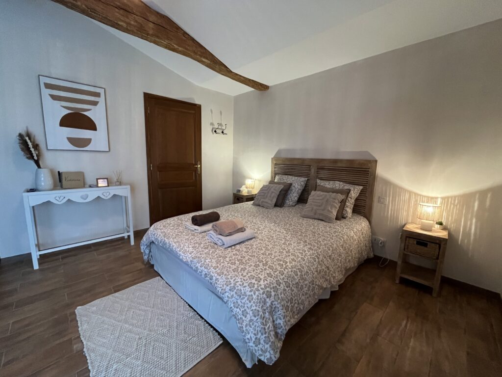Une chambre à coucher confortable avec un lit double, une literie à motifs et deux tables de chevet avec lampes. Une porte en bois, une console décorative, un tableau mural et une poutre en bois apparente complètent cet espace chaleureux et accueillant.
