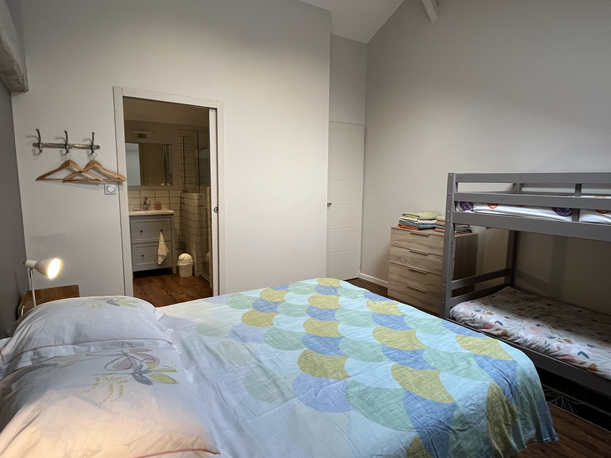 Une chambre confortable avec un lit double, un lit superposé, une commode en bois avec des serviettes pliées, des crochets muraux avec des cintres, une lampe de chevet, et une porte ouverte menant à une salle de bains avec un lavabo et un miroir.