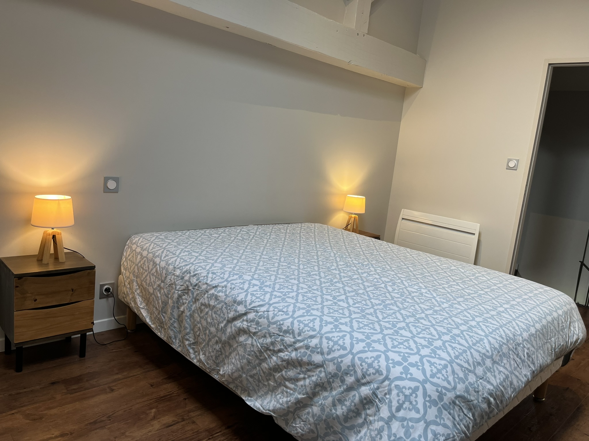 Un lit soigneusement fait avec un couvre-lit à motifs bleu clair et blanc, deux tables de nuit en bois avec des lampes allumées de chaque côté, et du parquet dans une chambre à la lumière tamisée.