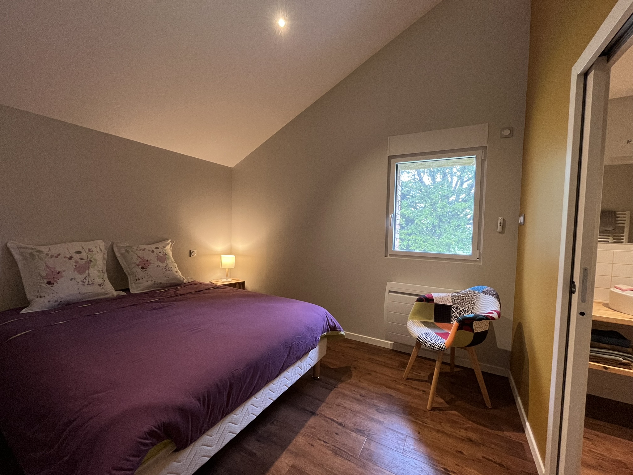 Une chambre à coucher confortable avec un lit double recouvert d'une couette violette, deux oreillers fleuris, une petite lampe de chevet, une chaise colorée près d'une fenêtre et du parquet. Des arbres sont visibles à travers la fenêtre.