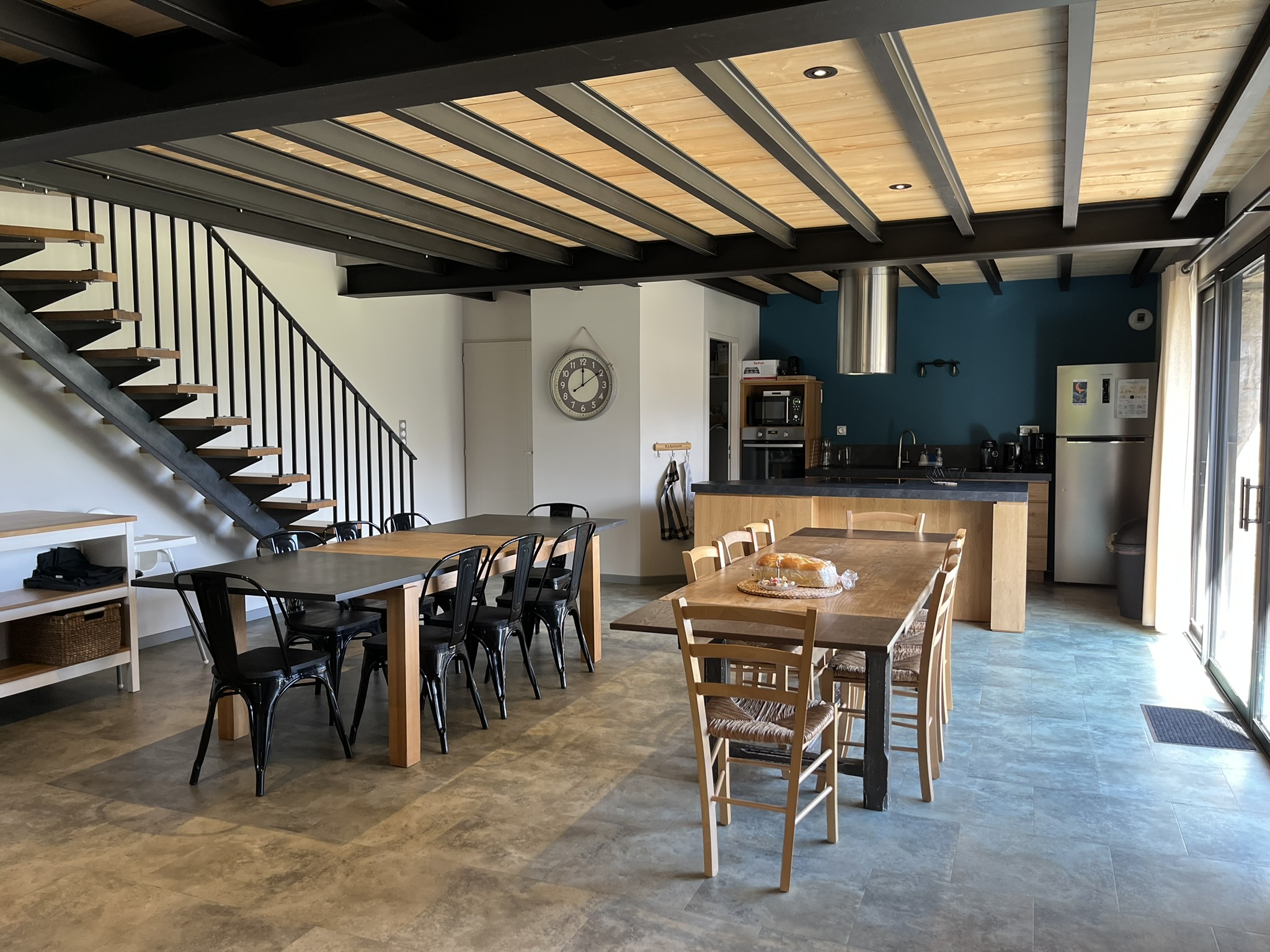 Cuisine et salle à manger modernes à aire ouverte avec des tables en bois, des chaises noires, un escalier sur la gauche et une cuisine avec des appareils électroménagers contre un mur d'accent sarcelle. De grandes fenêtres apportent la lumière naturelle.