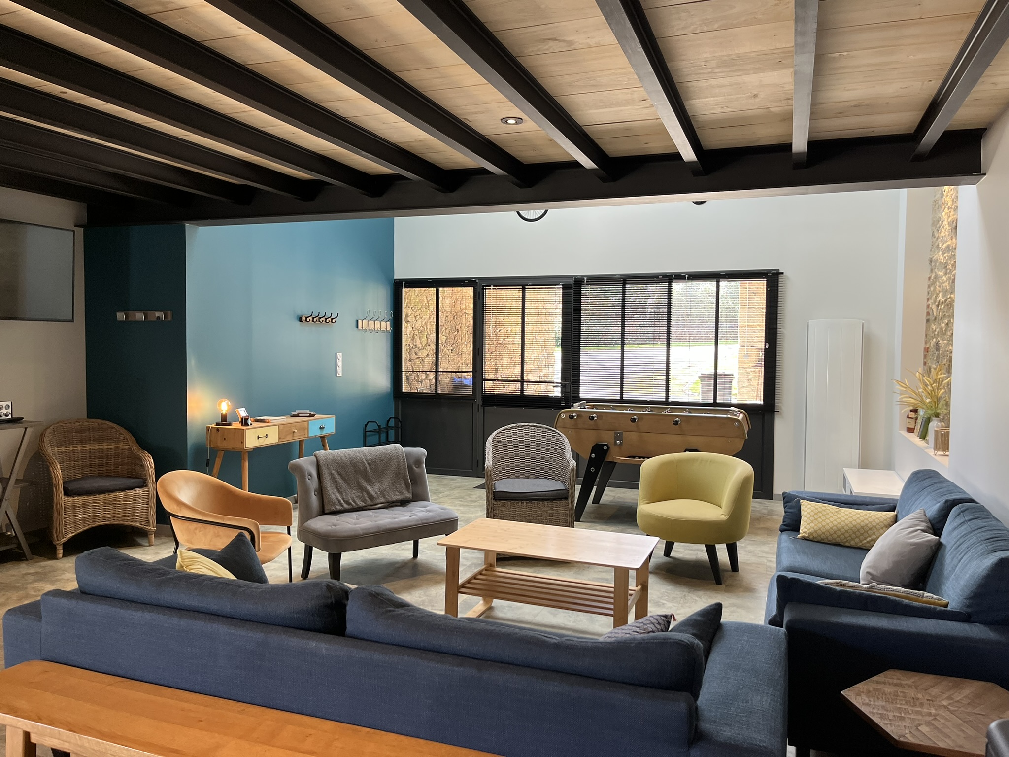 Un salon confortable avec des canapés, des fauteuils, une table basse en bois, un baby-foot près de grandes fenêtres et un mur d'accent sarcelle. Un éclairage chaleureux et un décor moderne et décontracté créent une atmosphère accueillante.