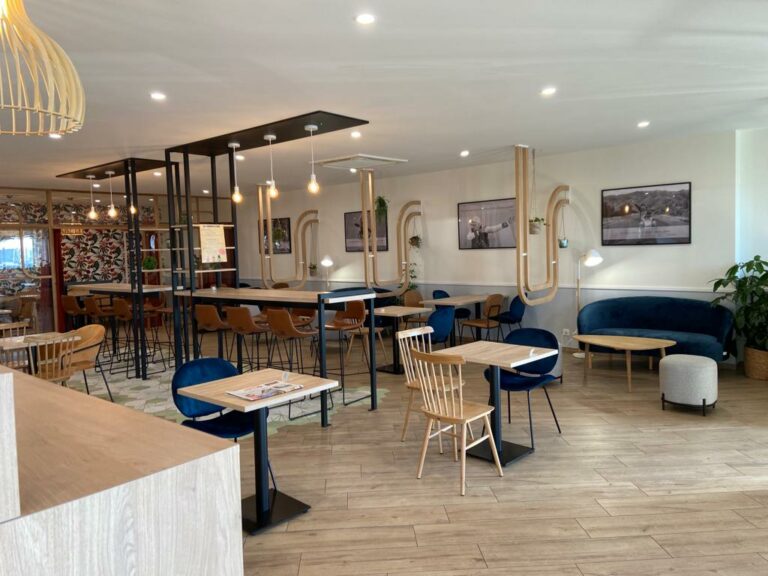 Un intérieur de café moderne avec des sols en bois et des meubles en bois clair, des tables, des chaises, des sièges à coussins bleus, des lampes suspendues, des œuvres d'art murales et des plantes - proche du Puy du Fou.