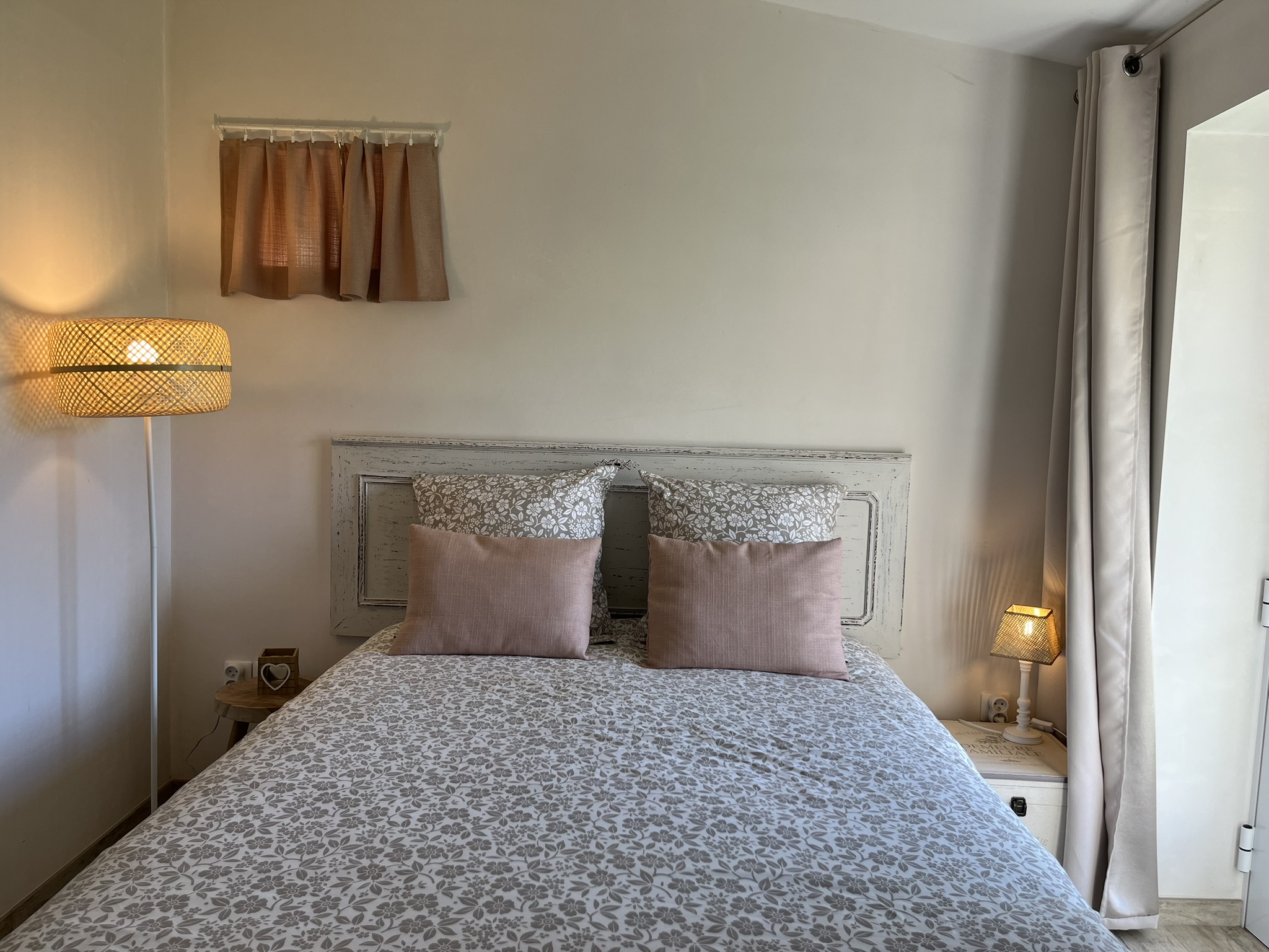 Un lit bien fait avec un couvre-lit gris et blanc à motifs, deux grands coussins fleuris et deux coussins d'appoint roses. Il y a de petites lampes de chaque côté et une minuscule fenêtre à rideaux au-dessus du lit. La pièce bénéficie d'un éclairage doux et naturel.