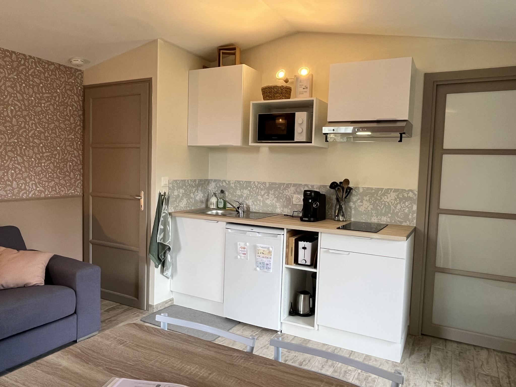 Une cuisine compacte et moderne comprend des armoires blanches, un micro-ondes, un évier, une cafetière, une plaque de cuisson électrique et un dosseret floral clair. À proximité se trouvent un canapé gris, une table en bois et des murs beiges à motifs.