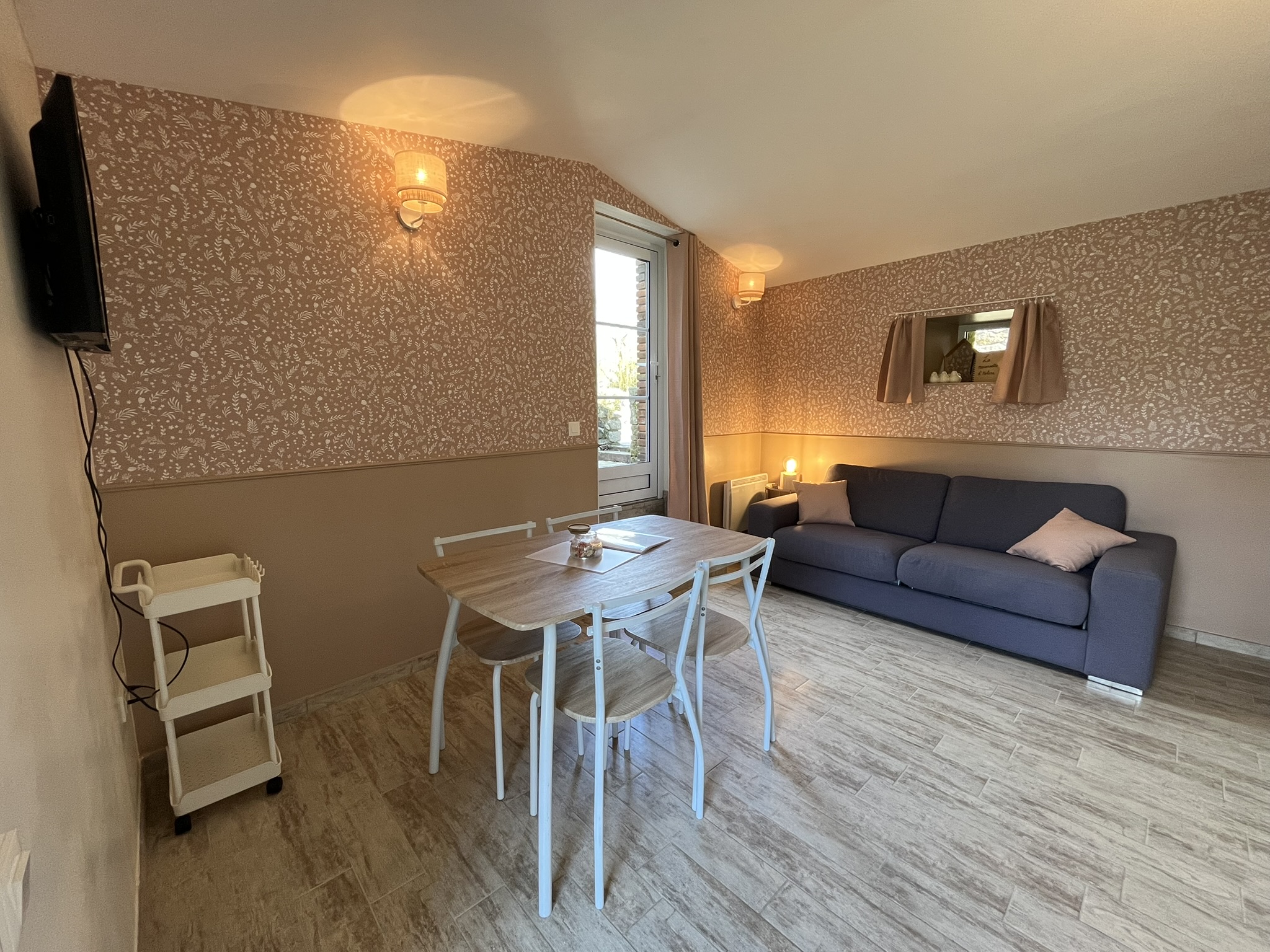 Un salon et une salle à manger confortables avec une table en bois et quatre chaises, un canapé bleu avec un coussin rose, une télévision murale et un papier peint à motifs. La lumière du soleil pénètre par une fenêtre et illumine l'espace.