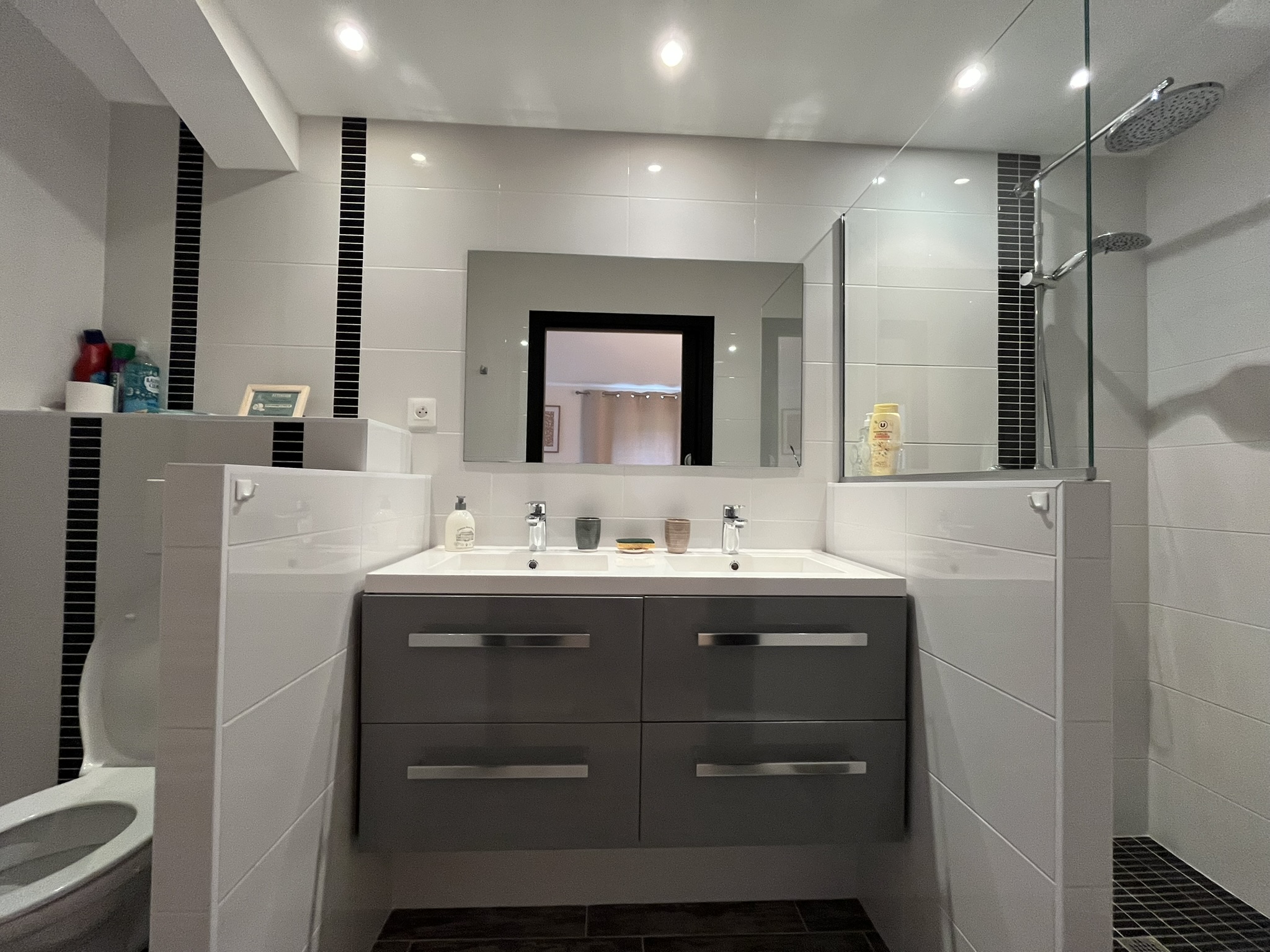 Salle de bains moderne avec un meuble à double vasque, un grand miroir et une douche en verre. Les articles de toilette sont placés sur des étagères et sur le lavabo. L'espace comporte des carreaux blancs avec des accents verticaux noirs et un éclairage zénithal lumineux.