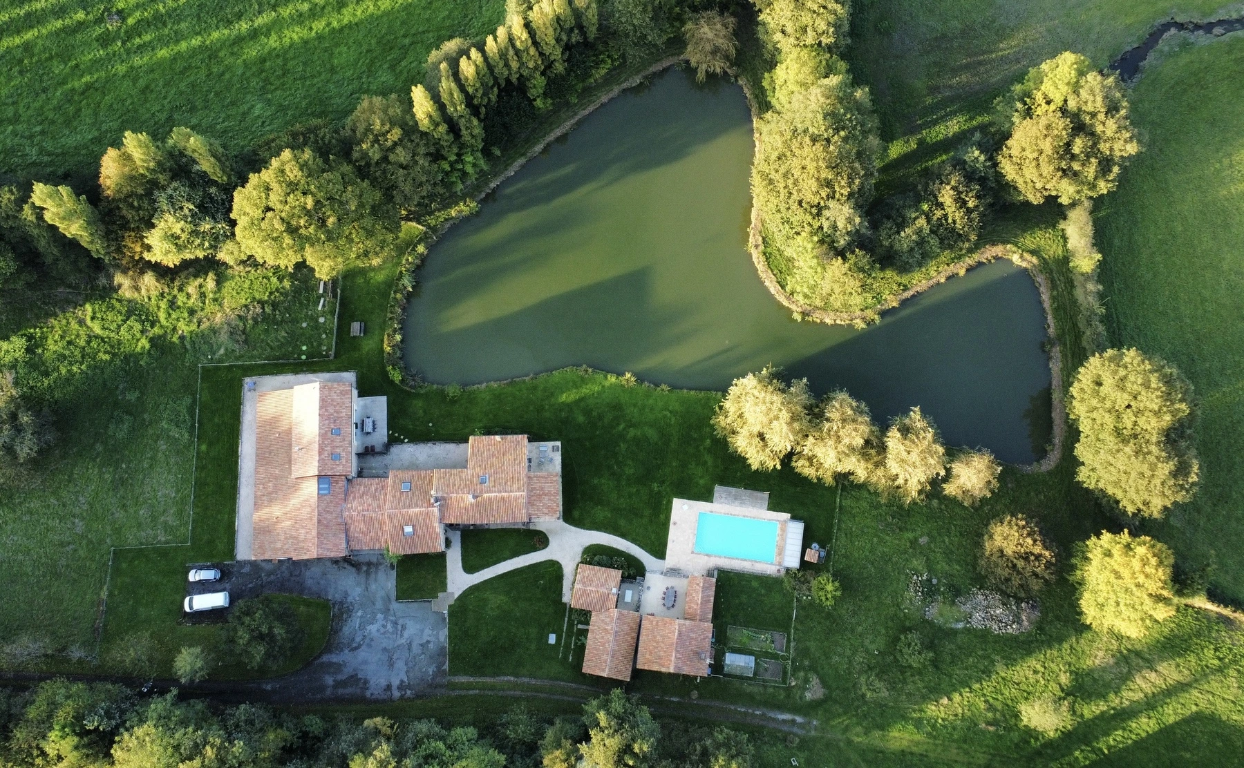 Un panneau en bois portant l'inscription L'ETANG NEUF pointe vers la gauche à partir d'un arbre à côté d'un champ herbeux, avec une clôture grillagée, des arbres et des bâtiments aux toits rouges visibles sous un ciel bleu clair.