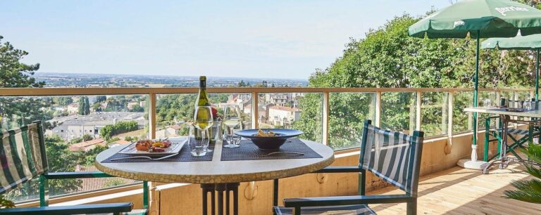 Un balcon parfait pour un séjour ou des vacances reposantes, avec une table ronde pour deux, des repas et du vin, donne sur une vue pittoresque de la ville. Des chaises en bandes et un parasol vert complètent cet hébergement accueillant sous un ciel clair.