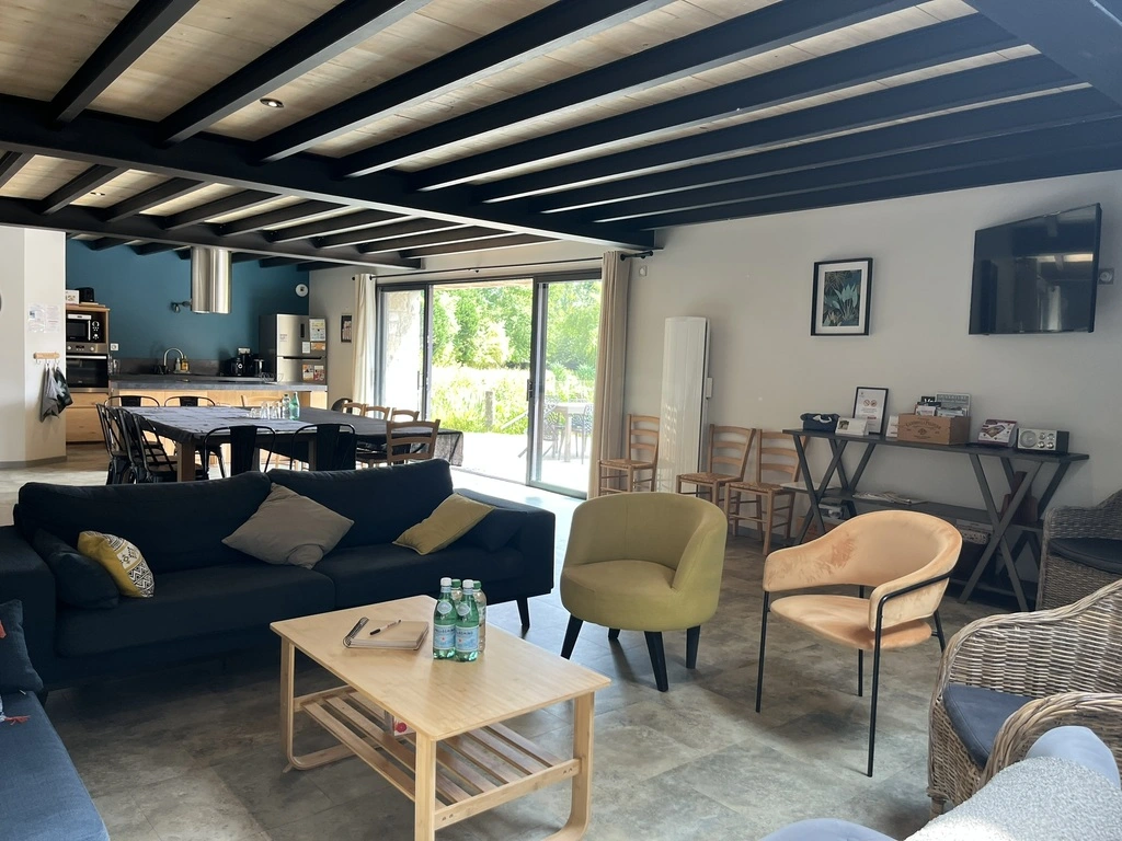 Un salon et une salle à manger spacieux et ensoleillés avec des canapés, des fauteuils, une table basse en bois, une table à manger avec des chaises et une cuisine ouverte. De grandes portes vitrées s'ouvrent sur la verdure à l'extérieur.