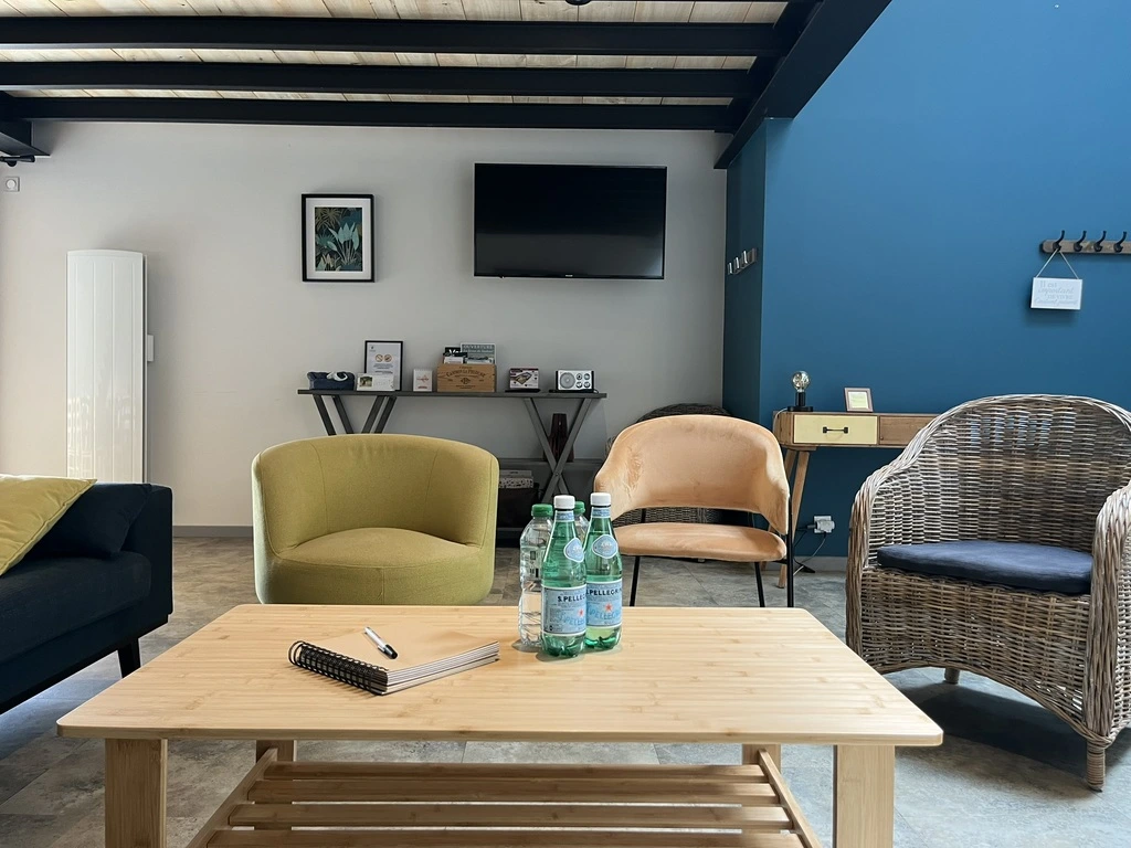Une salle d'attente confortable et moderne avec des chaises autour d'une table basse en bois sur laquelle sont posées des bouteilles d'eau et un carnet de notes ; un téléviseur est fixé au mur, et la pièce est ornée d'un mur d'accent bleu et d'œuvres d'art encadrées.