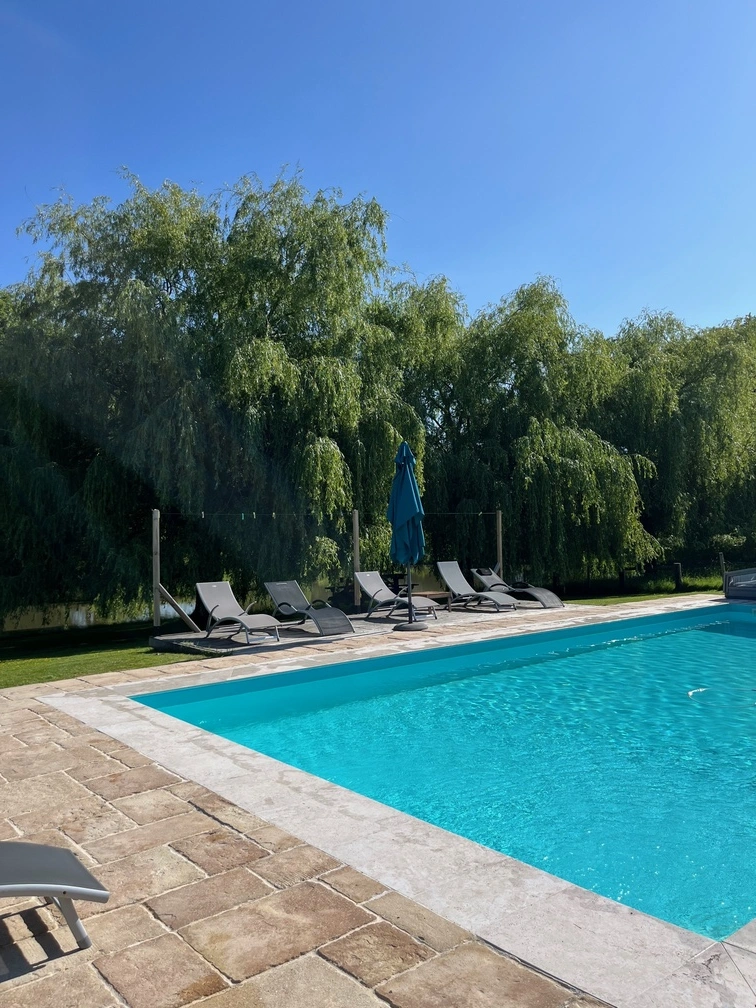 Une piscine à l'eau bleue claire est entourée de carreaux de pierre. Plusieurs chaises longues et un parasol bleu fermé se trouvent à côté de la piscine. Des arbres verdoyants bordent l'arrière-plan sous un ciel bleu clair.