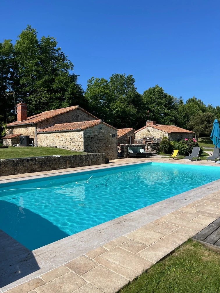 Au premier plan, une piscine bleue claire avec des carreaux de pierre est entourée d'herbe. Derrière la piscine se trouvent des maisons rustiques en pierre avec des toits en tuiles rouges, des arbres, des chaises longues et un parasol bleu sous un ciel d'un bleu éclatant.