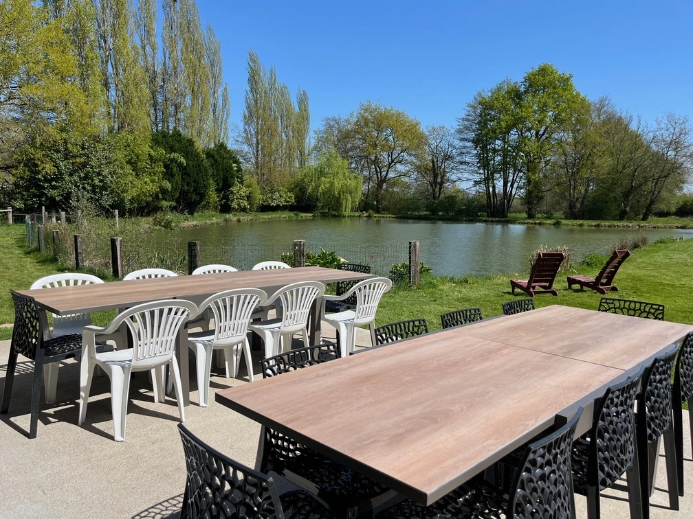 Plusieurs longues tables avec des chaises sont installées à l'extérieur, sur un patio donnant sur une pelouse et un petit étang entouré d'arbres, sous un ciel bleu limpide.