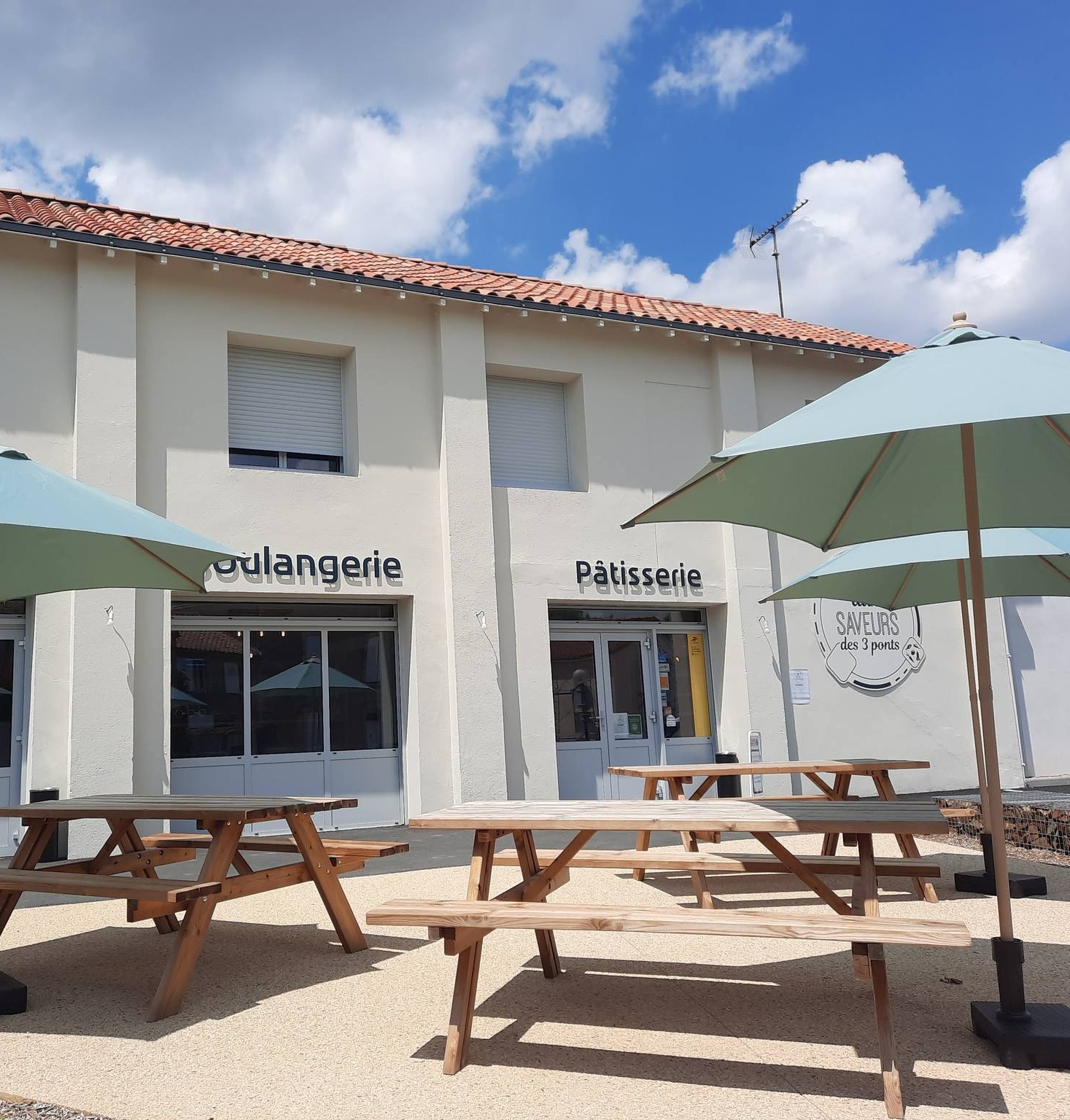Espace extérieur avec tables de pique-nique en bois et grands parasols devant un bâtiment blanc étiqueté boulangerie et pâtisserie, idéal pour un séjour de détente ou des vacances sous un ciel partiellement nuageux.