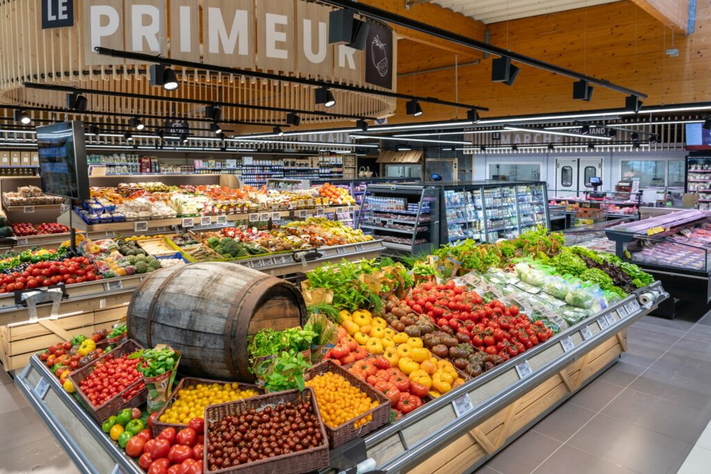 Rayon des produits frais dans un supermarché avec des fruits et légumes colorés bien rangés, rappelant un marché que vous pourriez visiter pendant votre séjour ou votre lieu de vacances, exposés autour d'un tonneau en bois sous un panneau indiquant LE PRIMEUR.