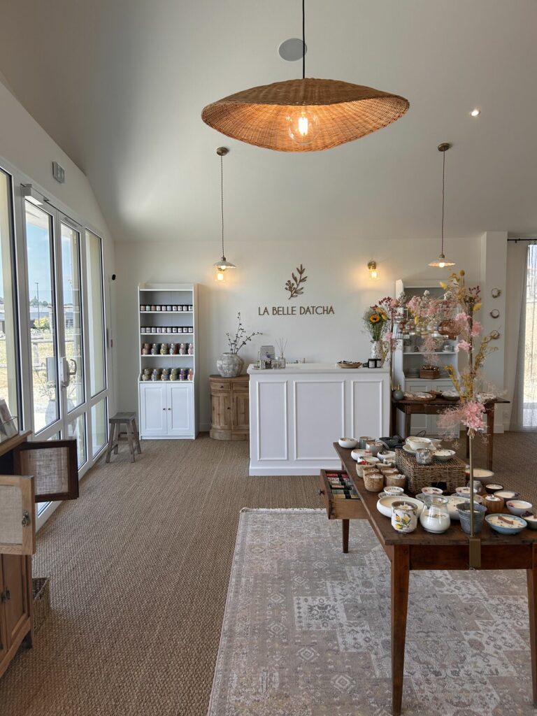 Une boutique lumineuse et accueillante avec un comptoir blanc, des étagères de bocaux et des tables présentant des céramiques et des objets de décoration inspirés par les alentours. Une douce lumière naturelle traverse les grandes fenêtres sur la gauche, éclairant un grand luminaire tissé.