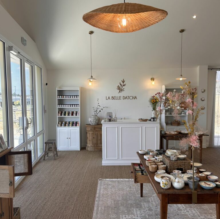 Une boutique lumineuse et accueillante avec un comptoir blanc, des étagères de bocaux et des tables présentant des céramiques et des objets de décoration inspirés par les alentours. Une douce lumière naturelle traverse les grandes fenêtres sur la gauche, éclairant un grand luminaire tissé.
