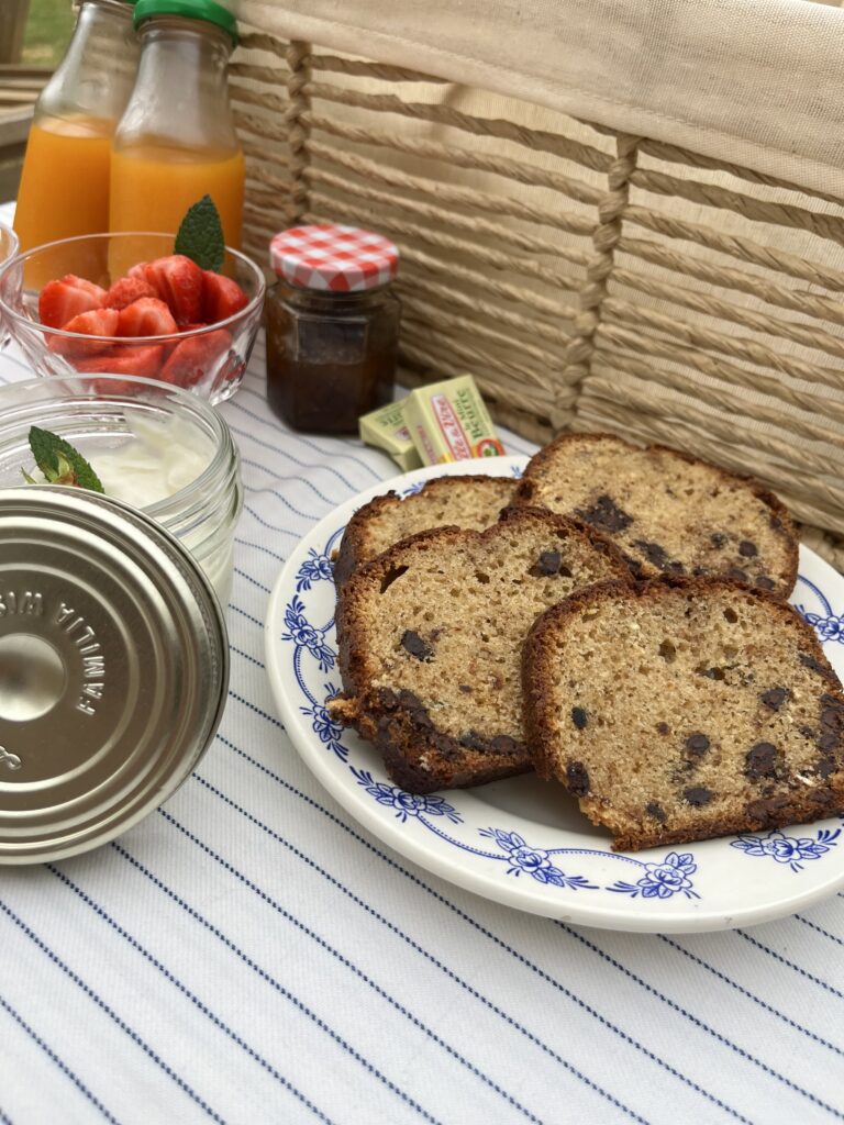 Un petit-déjeuner composé de tranches de pain de banane avec des pépites de chocolat sur une assiette fleurie, un yaourt aux fraises avec des fraises fraîches, deux bouteilles de jus, un pot de confiture et des sachets de beurre - parfaits pour profiter des hébergements et des offres spéciales.
