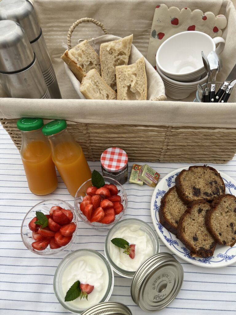 Un délicieux pique-nique sur une nappe rayée comprend du pain dans un panier, des tasses, des couverts, deux bouteilles de jus d'orange, des bols de fraises et de yaourt, des tranches de pain aux fruits et des mini-pots de confiture et de beurre - parfaits pour déguster les offres exclusives des hébergements locaux.