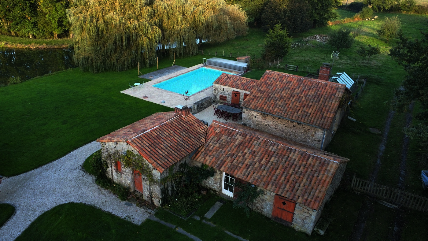 Vue aérienne de chalets rustiques en pierre aux toits de tuiles rouges, entourés d'herbe verte et d'arbres, avec une piscine bleue et un patio dans l'arrière-cour.