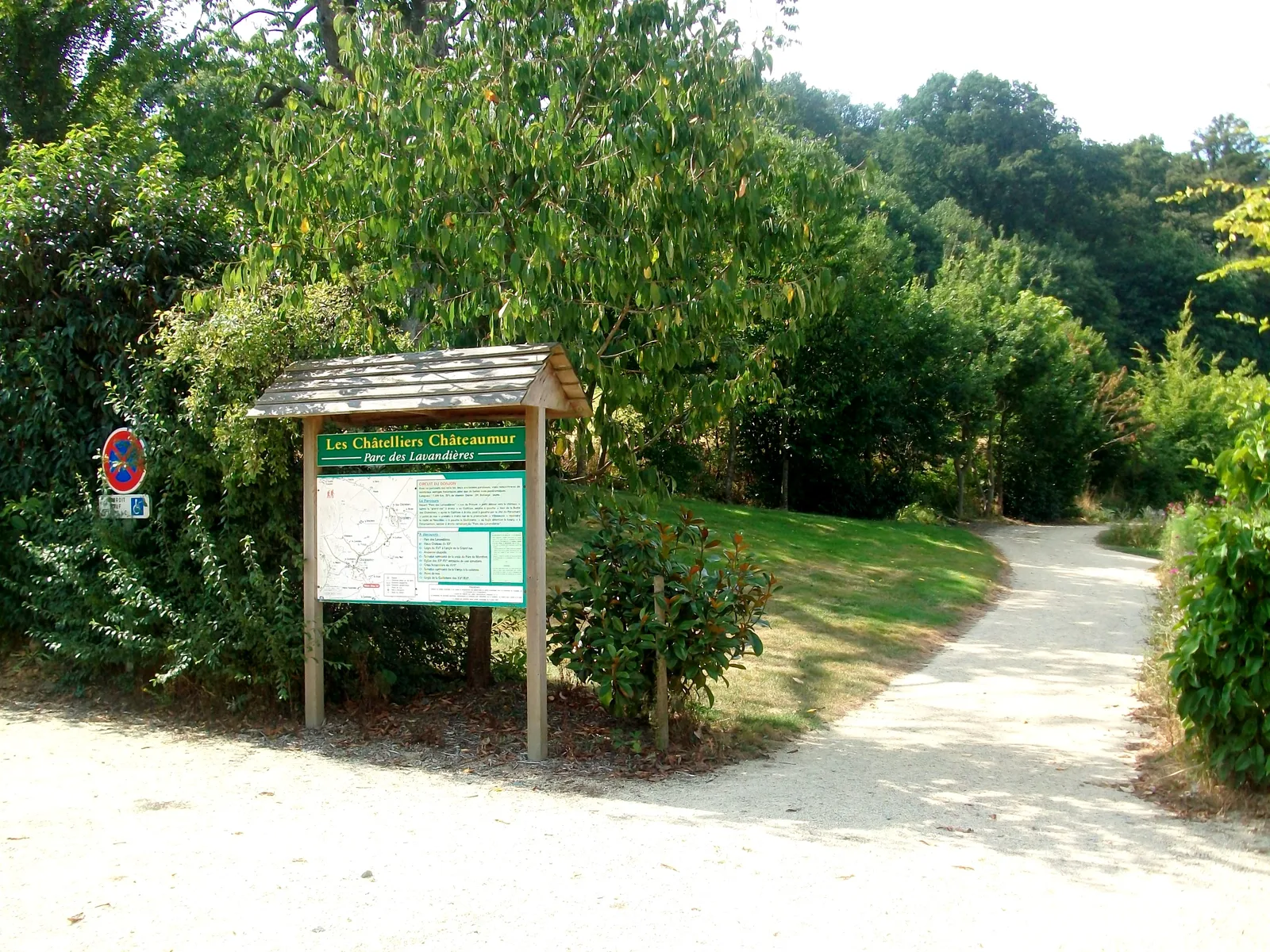 Une entrée de sentier du parc avec un panneau d'information en bois et un petit panneau sur la gauche, entourée d'arbres verts et de buissons aux alentours, avec un chemin de gravier incurvé qui disparaît dans le feuillage.