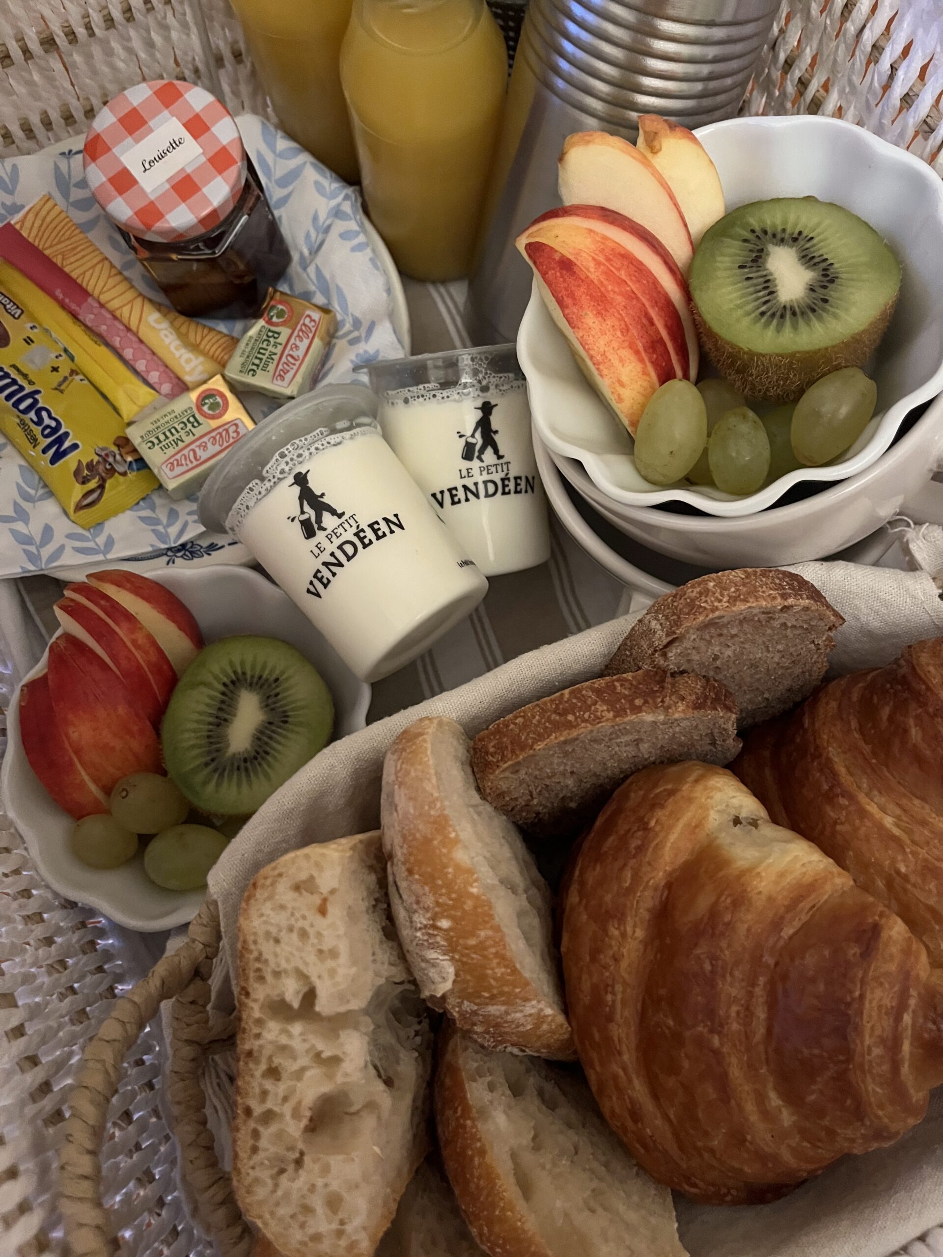 Un délicieux pique-nique sur une nappe rayée comprend du pain dans un panier, des tasses, des couverts, deux bouteilles de jus d'orange, des bols de fraises et de yaourt, des tranches de pain aux fruits et des mini-pots de confiture et de beurre - parfaits pour déguster les offres exclusives des hébergements locaux.