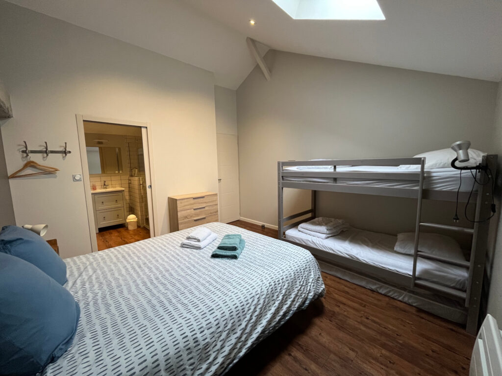La chambre confortable de nos gîtes de Vendée comprend un lit double, un lit superposé, une commode et une salle de bains privative. Les serviettes sont pliées sur le lit et les planchers en bois ajoutent de la chaleur.