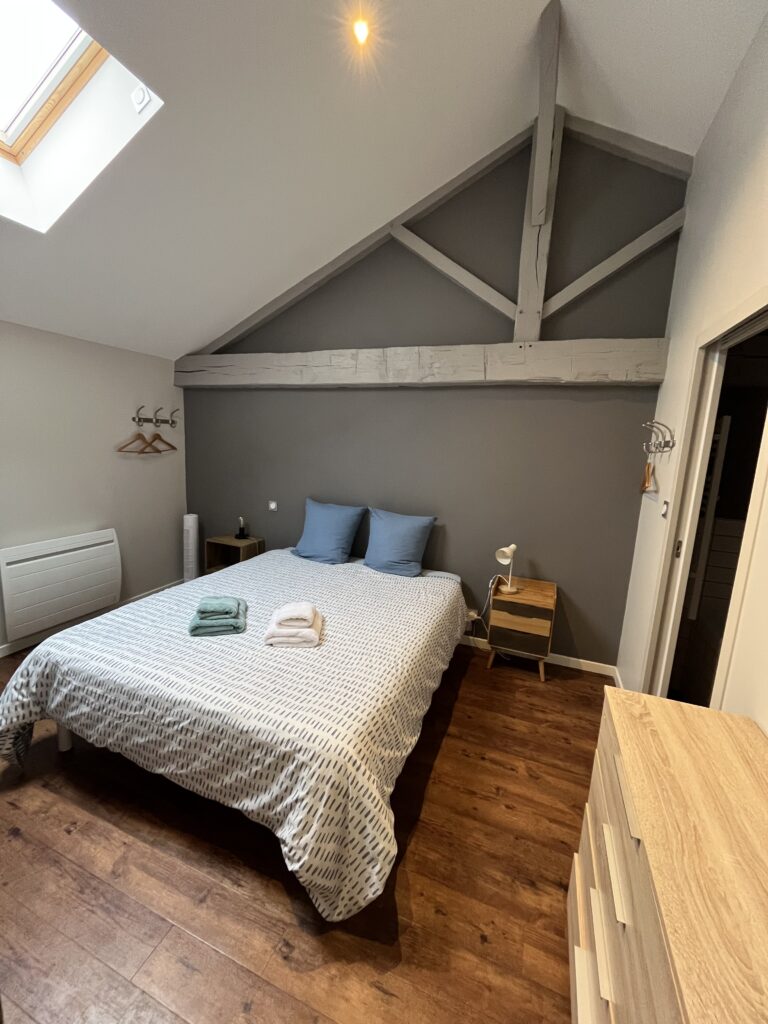 Un lit double bien fait avec des serviettes pliées, des tables de nuit et des poutres au plafond incliné dans une chambre moderne au sol en bois et aux murs neutres.