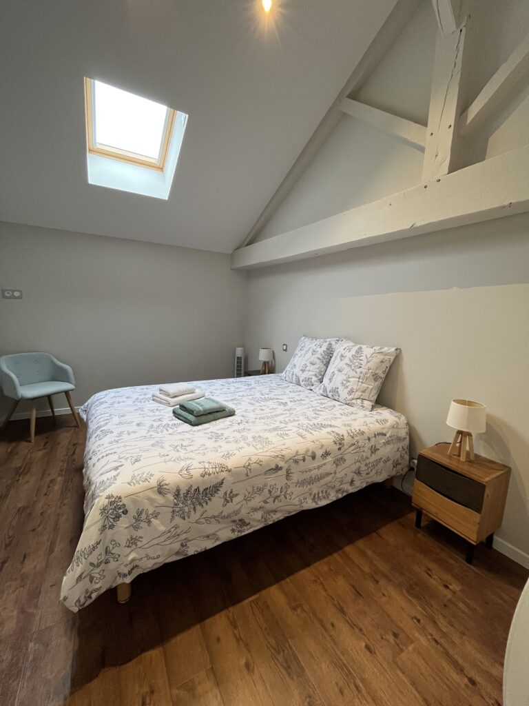 Un lit double soigneusement fait avec des draps à motifs, une table de chevet avec une lampe, une serviette pliée, une chaise bleue, un plancher en bois et une lucarne dans une chambre aux murs blancs.