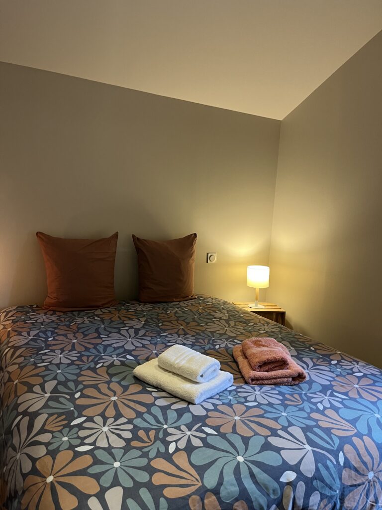 Un lit bien fait avec une couette à motifs floraux, deux oreillers bruns, des serviettes pliées sur le dessus, et une petite lampe de chevet allumée.