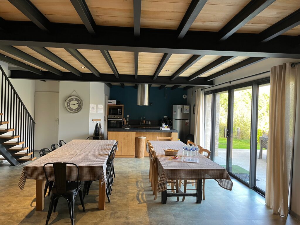 Salle à manger spacieuse avec deux tables recouvertes de nappes à carreaux, des chaises noires, une cuisine en arrière-plan et de grandes fenêtres donnant sur un espace vert en plein air.