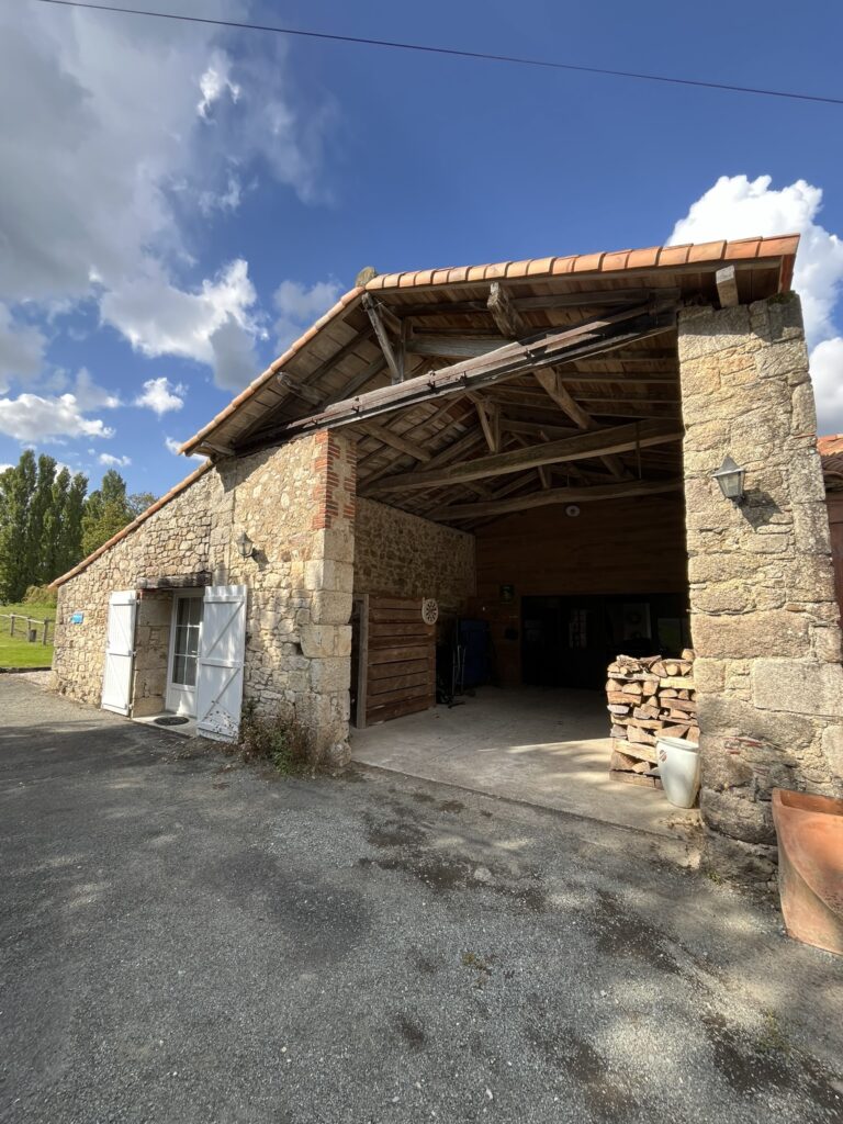 Grange en pierre avec toit en tuiles, entrée ouverte, bois de chauffage empilé, portes blanches et allée en gravier sous un ciel partiellement nuageux.