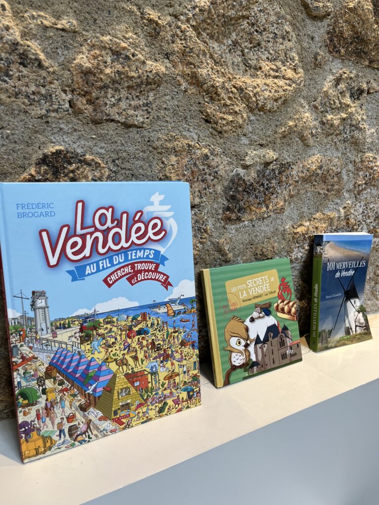 Trois livres en français sur la région de la Vendée sont exposés verticalement sur une étagère blanche contre un mur en pierre texturée.
