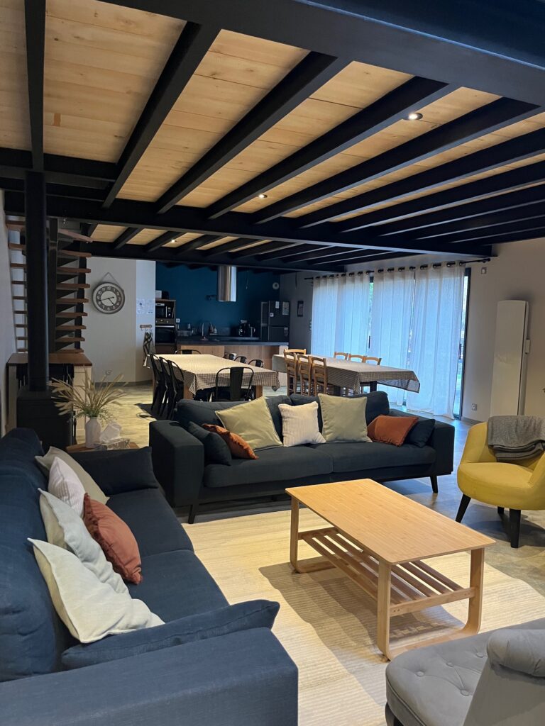Salon moderne à aire ouverte avec deux canapés, une table basse en bois, un coin repas et une cuisine visible, avec des poutres apparentes et de grandes fenêtres avec des rideaux à voilage.