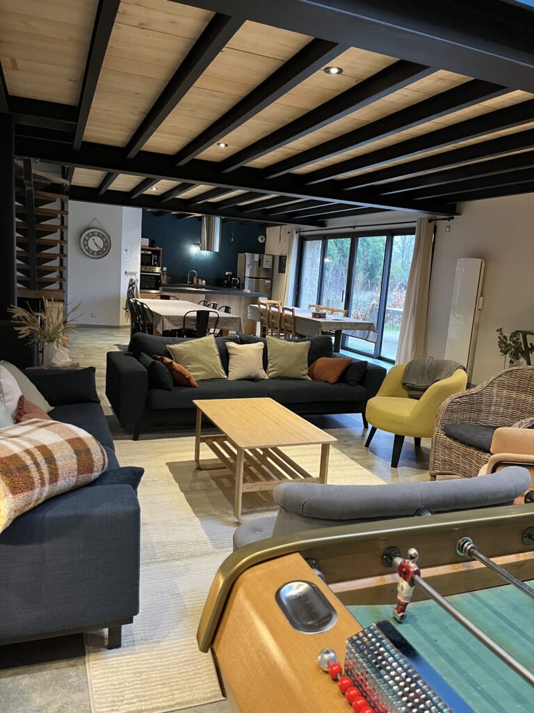 Espace de vie ouvert et moderne avec canapés, table basse, baby-foot, table à manger et cuisine en arrière-plan. De grandes fenêtres laissent entrer la lumière naturelle.