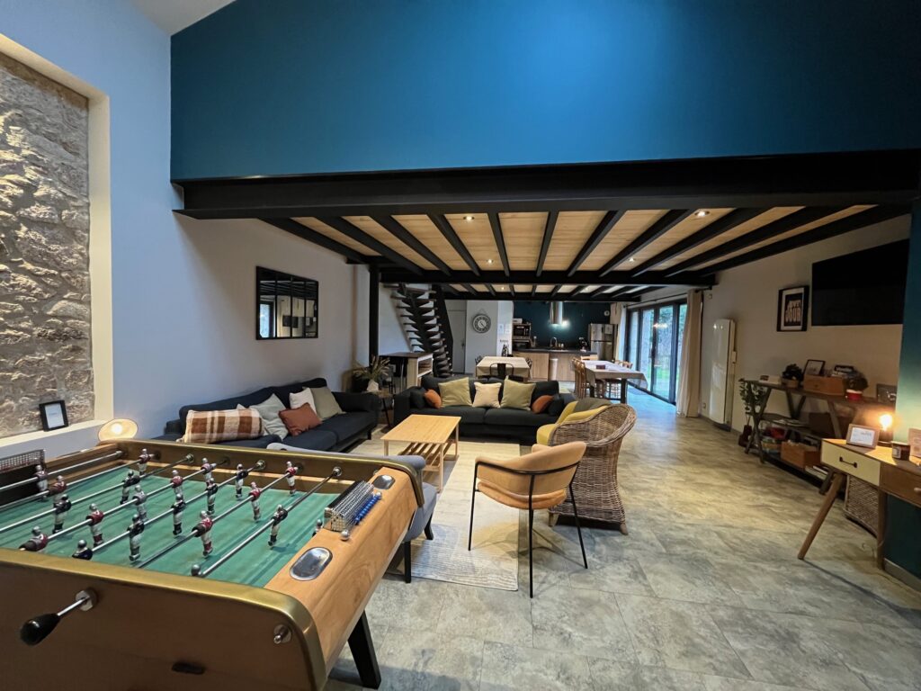 Salon moderne et spacieux avec un baby-foot au premier plan, des canapés, un coin repas et de grandes fenêtres apportant de la lumière naturelle.