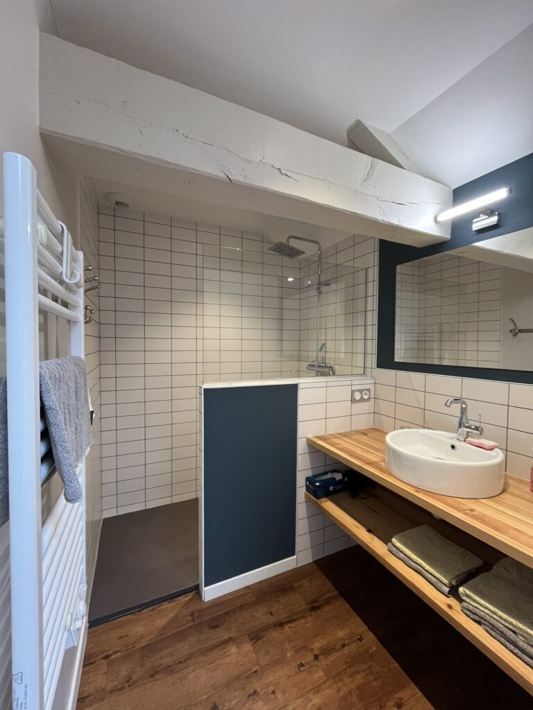 Salle de bains moderne avec douche à l'italienne, murs carrelés en blanc, plan de travail en bois avec lavabo, grand miroir, porte-serviettes et serviettes pliées sur des étagères.