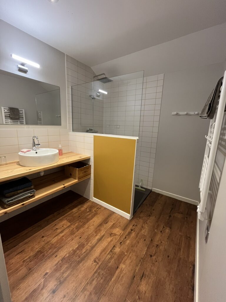 Salle de bains moderne avec sol en bois, meuble-lavabo flottant en bois, vasque, miroir sans cadre, douche à l'italienne avec panneau en verre et cloison jaune. Les serviettes sont sur l'étagère et le radiateur.