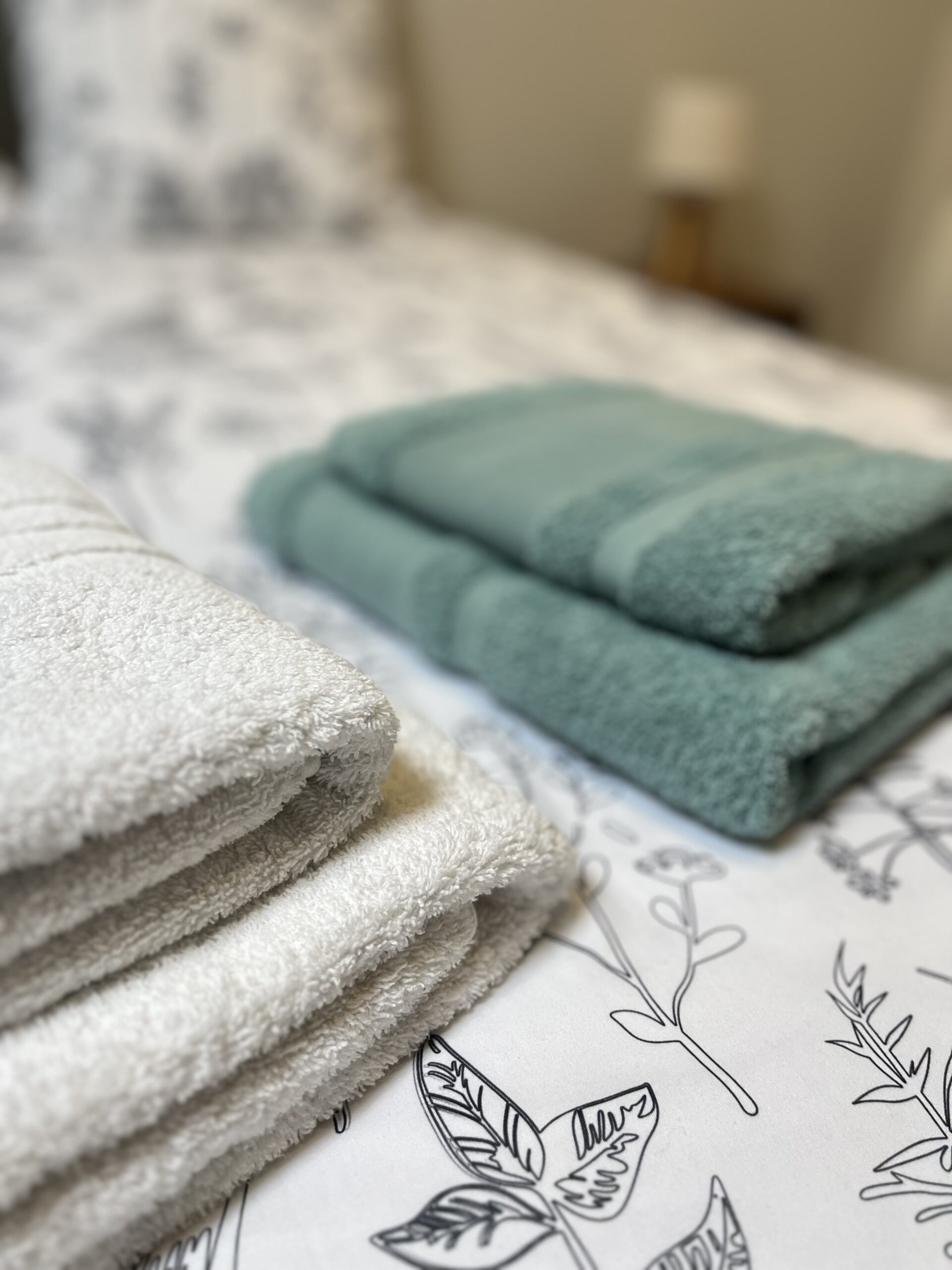 Deux serviettes soigneusement pliées, l'une blanche et l'autre verte, posées sur un lit au couvre-lit à motifs floraux.