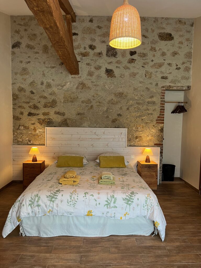 Une chambre confortable avec un mur d'accent en pierre, un lit double avec une literie florale, deux oreillers jaunes, des tables de chevet avec des lampes et un luminaire suspendu en osier.