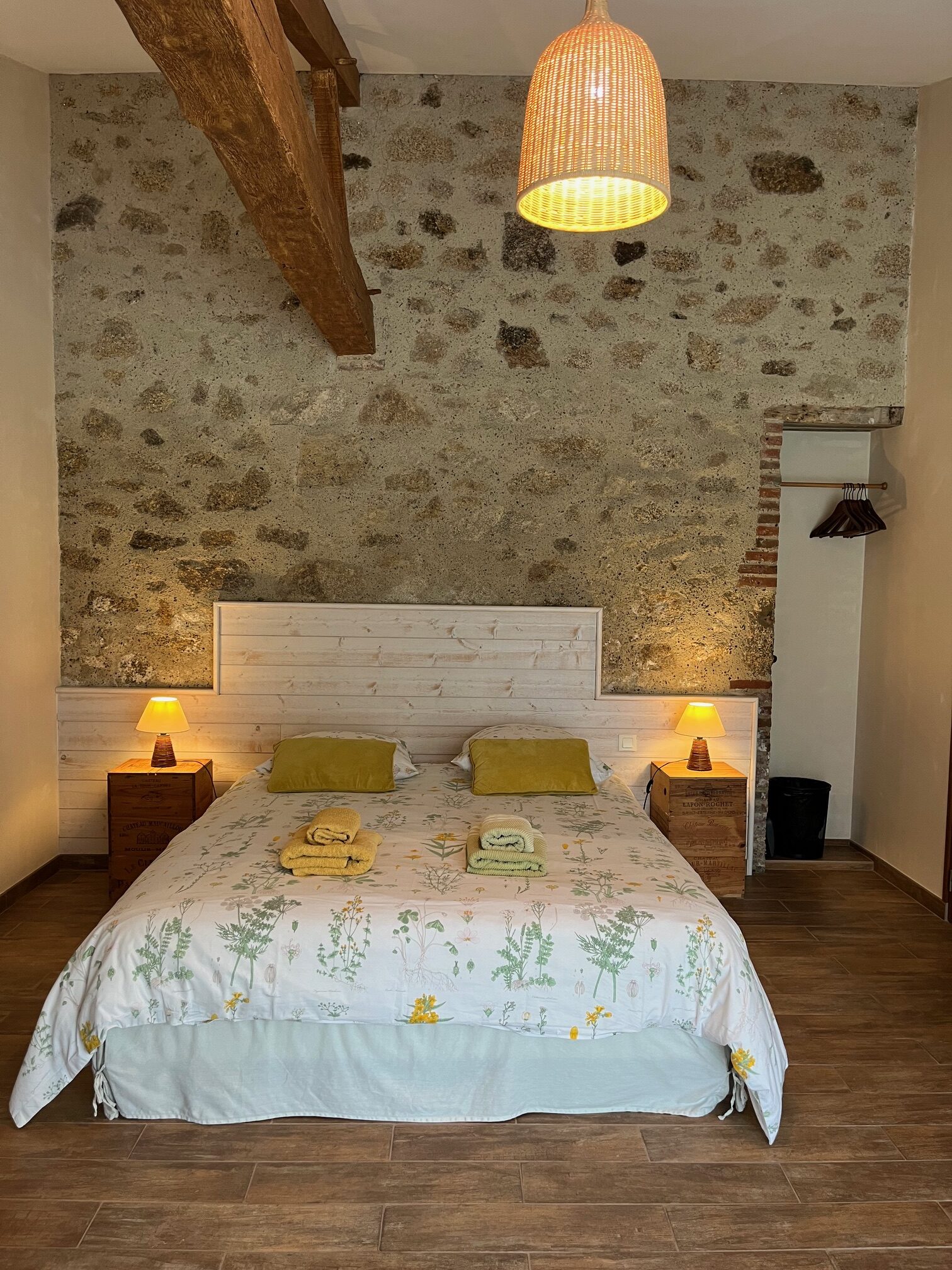 Une chambre confortable avec un mur d'accent en pierre, un lit double avec une literie florale, deux oreillers jaunes, des tables de chevet avec des lampes et un luminaire suspendu en osier.