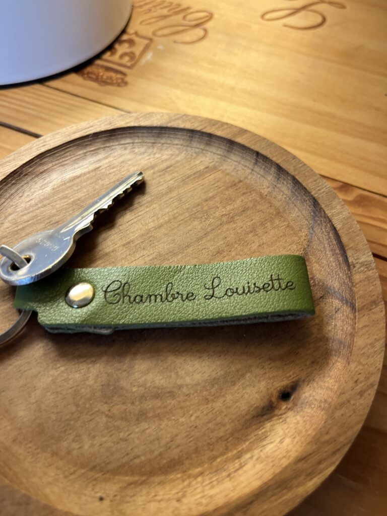 Une clé en argent attachée à un porte-clés en cuir vert avec l'emblème de la Chambre Louisotte, placée sur un plat rond en bois.