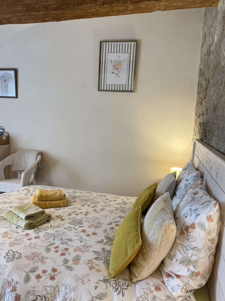 Un lit soigneusement fait avec des draps à fleurs, deux oreillers jaunes et des serviettes pliées, à côté d'une lampe allumée et d'une chaise en osier dans une chambre à coucher doucement éclairée.