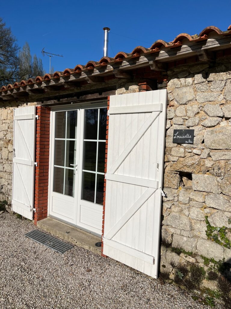 Bâtiment en pierre avec des doubles portes en verre encadrées de blanc et des volets blancs, sous un toit de tuiles rouges. Un panneau à côté de la porte indique "Louisette".