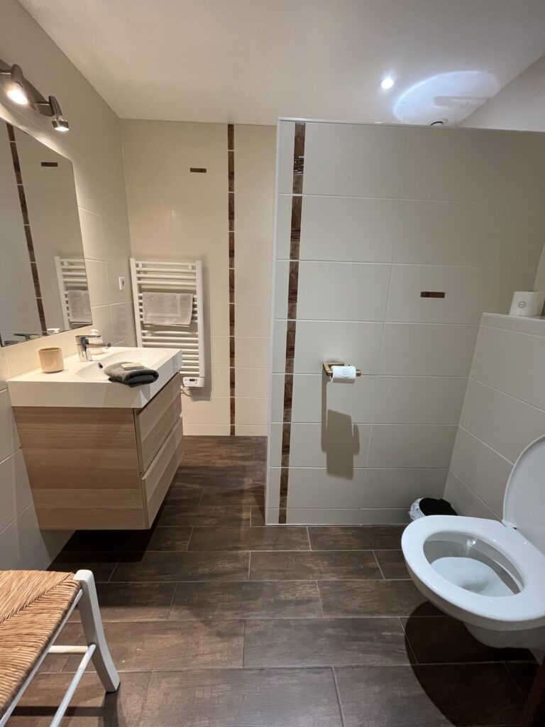 Salle de bains moderne avec des murs en carrelage beige, un sol en carrelage marron, des toilettes murales, un meuble avec un lavabo, un miroir, un sèche-serviettes et une chaise.