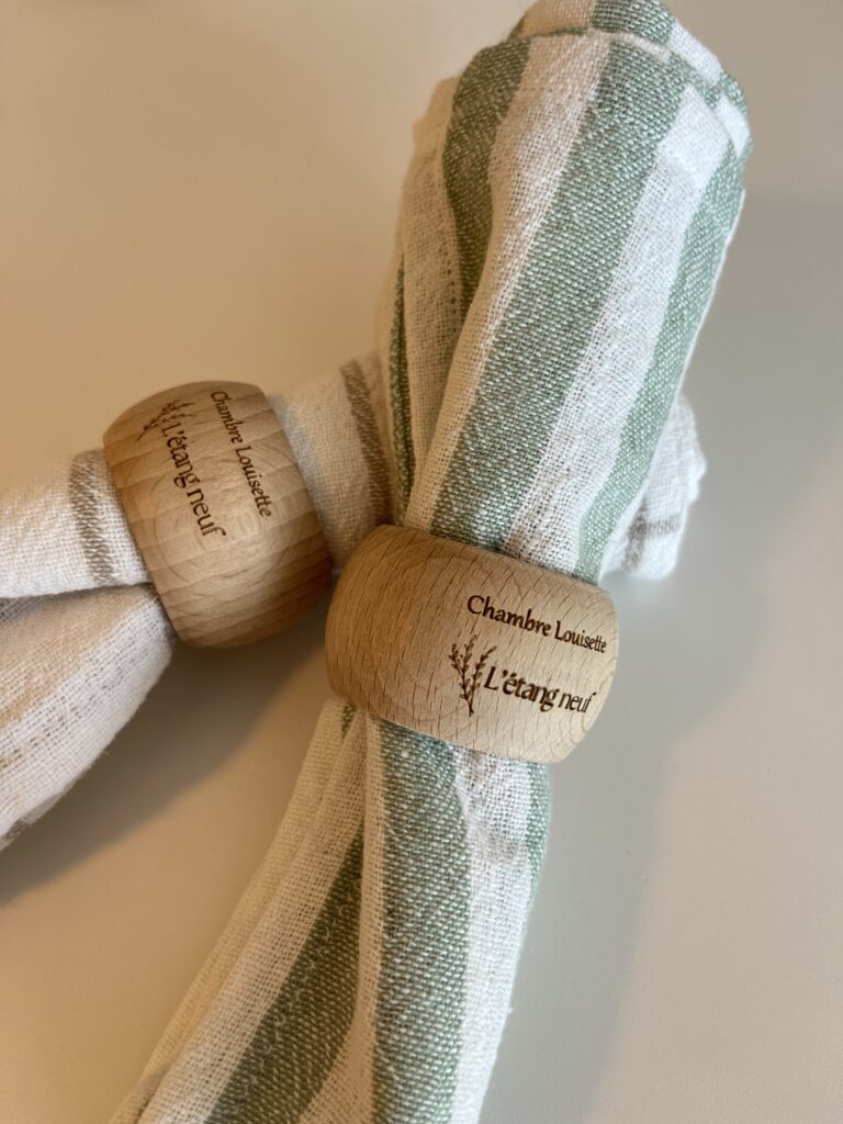 Deux serviettes en tissu rayé retenues par des ronds de serviette en bois gravés d'un texte et d'un petit motif végétal, posés sur une surface claire.