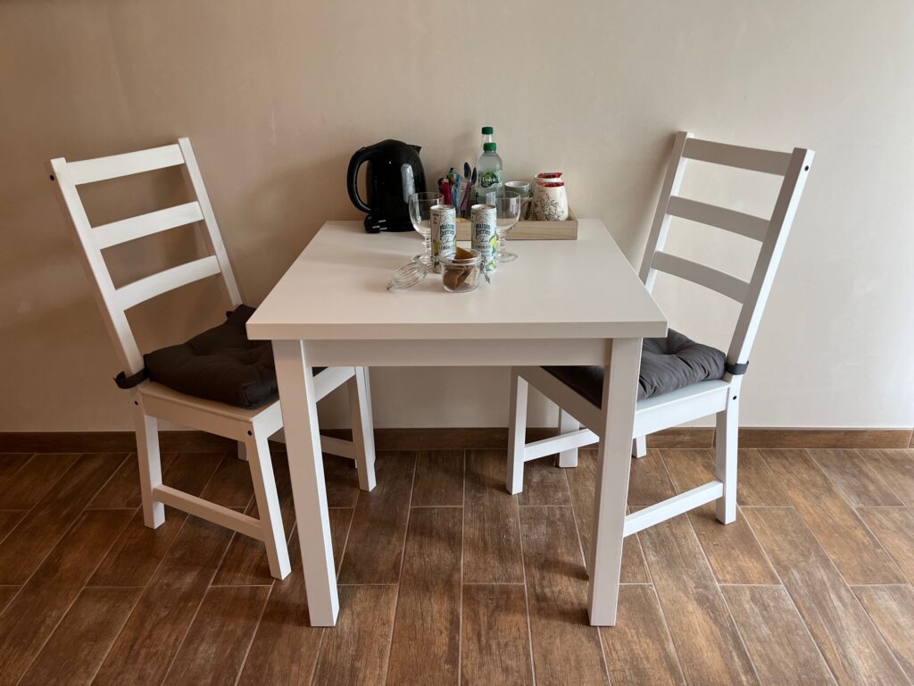 Une petite table blanche avec deux chaises blanches sur un sol en bois, avec une bouilloire, des bouteilles, des bocaux, des tasses et une assiette.