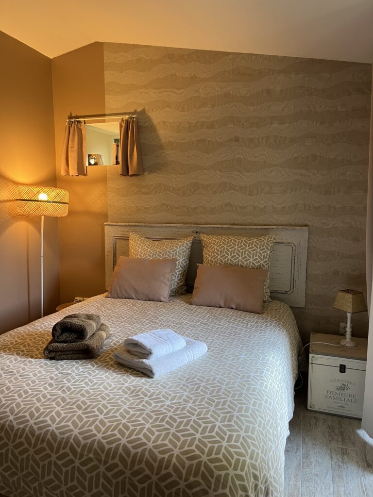 Un lit soigneusement fait avec des draps à motifs et des serviettes pliées sur le dessus, à côté d'une petite lampe et d'une table de nuit, dans une chambre à coucher aux tons beiges et à l'éclairage tamisé.