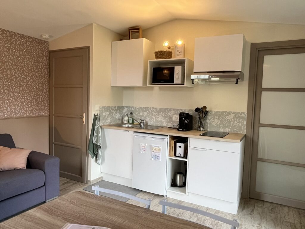 Kitchenette compacte avec armoires blanches, micro-ondes, cafetière, évier, bouilloire et plaque de cuisson, à côté d'une porte et d'un canapé dans une petite pièce bien éclairée.